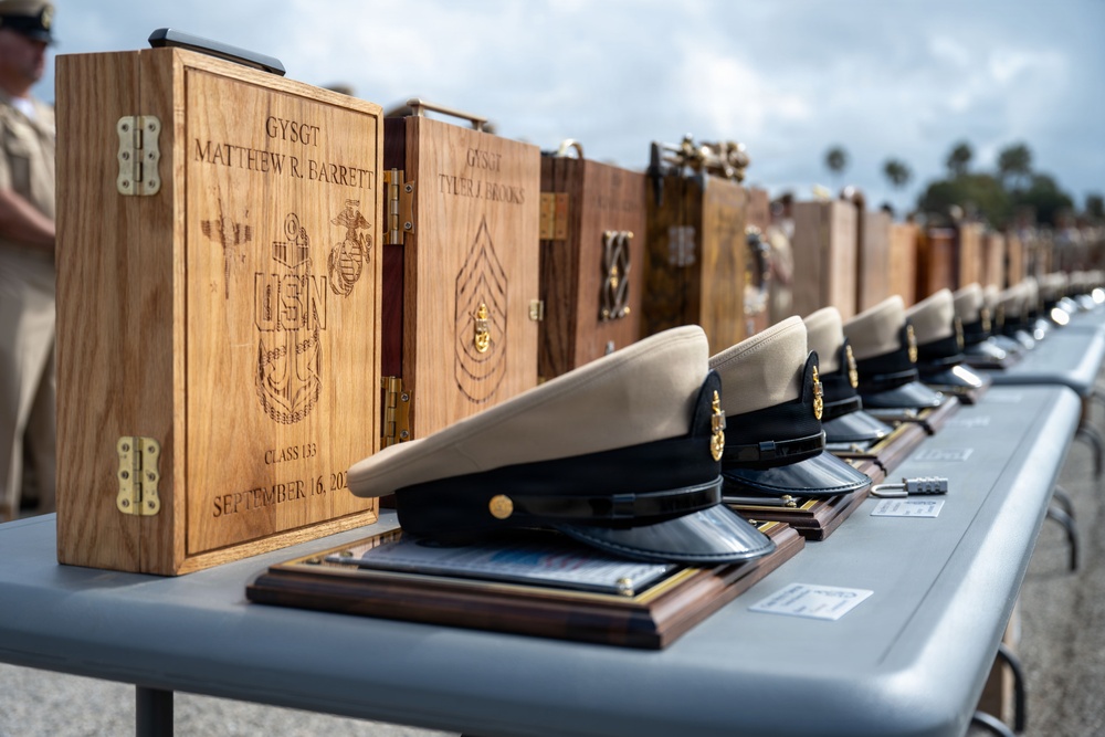 A formal military ceremony display showing a long table with engraved wooden keepsake boxes and dress uniform caps arranged in a row. The boxes are laser-engraved with USN and USMC insignia, individual names and ranks (GySgt), class number 133, and the date September 16. This appears to be a Navy Chief Petty Officer or joint military advancement/retirement ceremony.