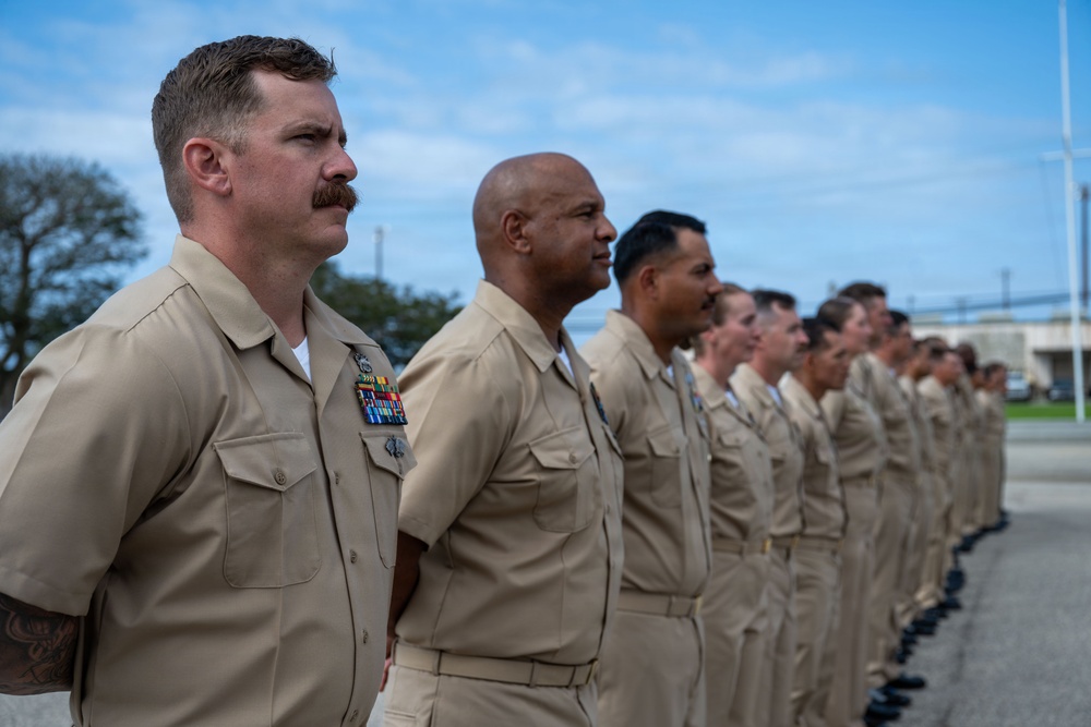 A formation of U.S. Navy personnel standing at attention in tan/khaki service uniform (Service Uniform Charlie). The group is arranged in a single diagonal line extending into the background, suggesting a formal military ceremony or inspection. The foreground sailor displays multiple service ribbons and awards on his uniform.
