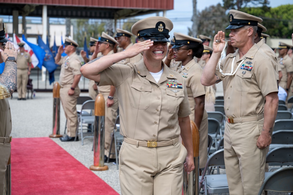 A formal U.S. Navy military ceremony is taking place outdoors, likely a change of command or retirement ceremony. A female senior enlisted sailor in Service Khaki uniform walks along a red carpet while rendering a salute, smiling broadly. Multiple Navy personnel are standing at attention and saluting in the background.