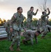 A group of U.S. Army soldiers in OCP (Operational Camouflage Pattern) uniforms are conducting physical training exercises outdoors at dawn or dusk. The soldiers are performing various movements including push-ups, squats, and standing exercises on a grassy area. The scene appears to be a structured physical training session at a military installation.