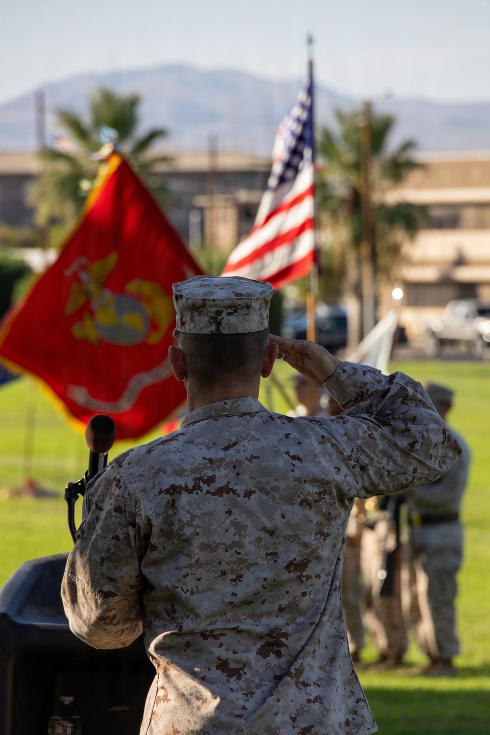 A U.S. Marine in desert MARPAT camouflage uniform stands at a podium rendering a salute during a formal military ceremony. The scene includes the U.S. Marine Corps flag and the American flag prominently displayed in the background. The setting appears to be an outdoor parade ground or ceremonial area at a military installation, likely in a desert region based on the terrain and palm trees visible.