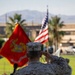 A U.S. Marine in desert MARPAT camouflage uniform stands at a podium rendering a salute during a formal military ceremony. The scene includes the U.S. Marine Corps flag and the American flag prominently displayed in the background. The setting appears to be an outdoor parade ground or ceremonial area at a military installation, likely in a desert region based on the terrain and palm trees visible.