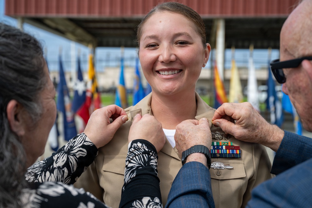A female U.S. Navy service member is smiling broadly during what appears to be a promotion or pinning ceremony. Two individuals, likely family members, are simultaneously pinning new rank insignia onto the collar of her tan/khaki Navy service uniform. Multiple military unit flags are visible in the background, indicating a formal military ceremony setting.