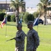 Two U.S. Marines in MARPAT desert camouflage uniforms stand at attention on a grass parade field during what appears to be a formal military ceremony. One Marine is holding a ceremonial baton or swagger stick. Flags and palm trees are visible in the background, suggesting a base in a warm, arid climate.