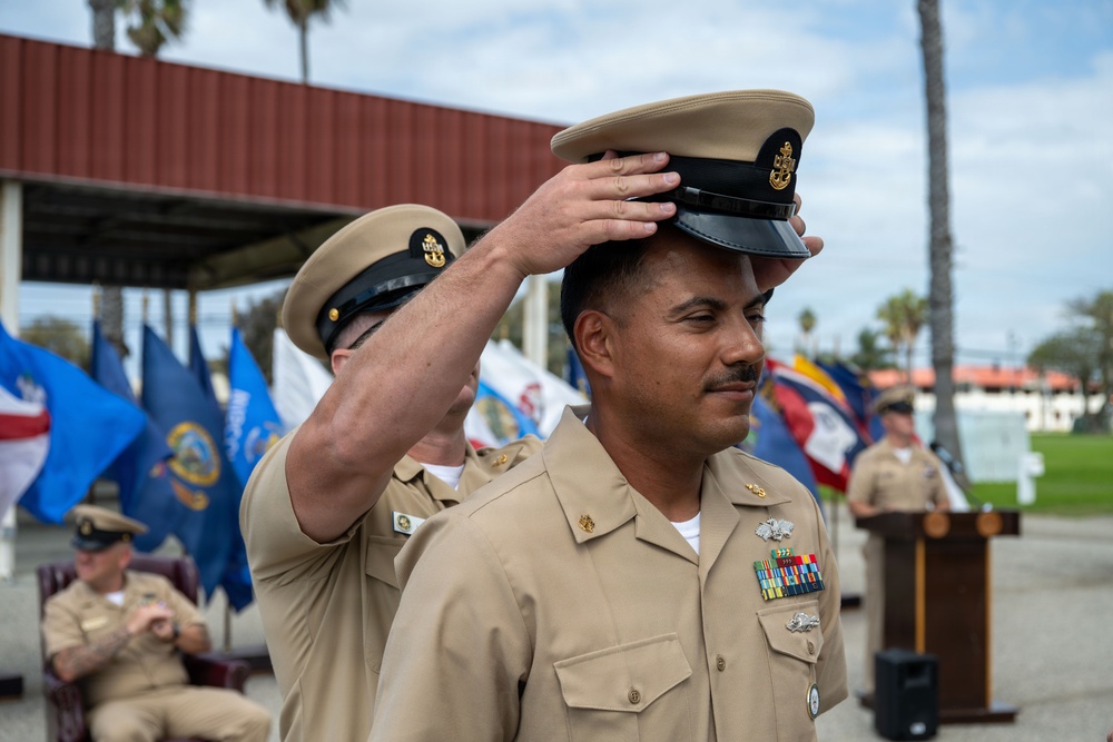 A U.S. Navy promotion or advancement ceremony is taking place outdoors at what appears to be a naval installation. One service member is placing a chief petty officer combination cover (hat) onto another sailor's head, a traditional part of the CPO pinning/advancement ceremony. Multiple military flags and personnel are visible in the background.