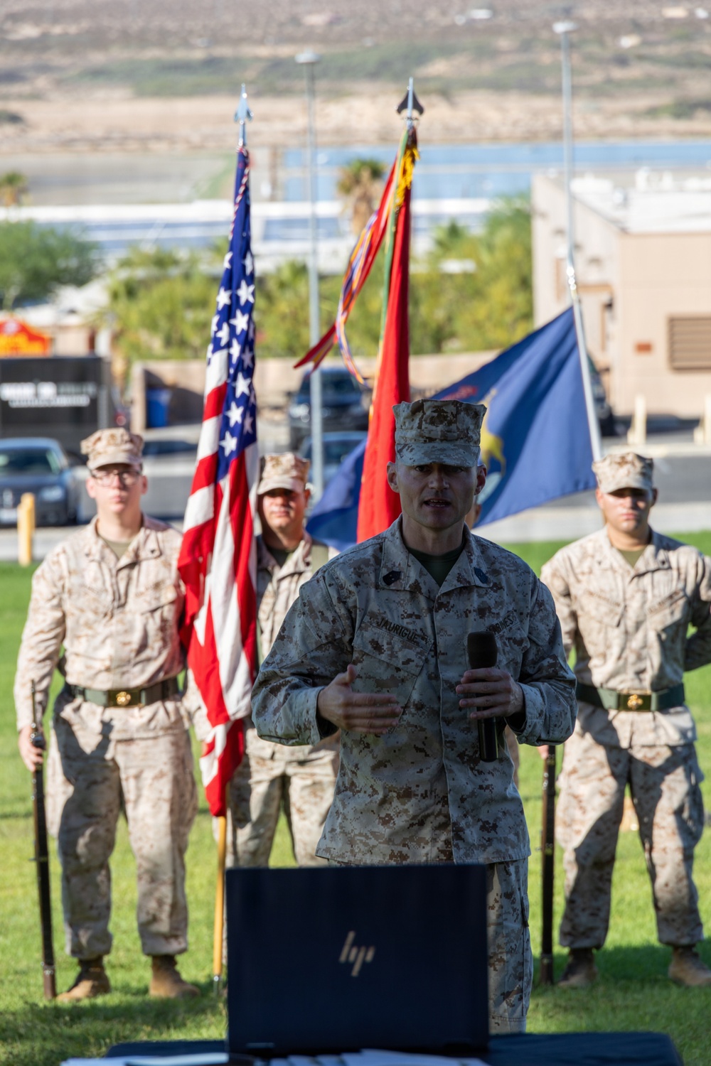 A U.S. Marine in desert MARPAT camouflage uniform is speaking at a podium holding a microphone during what appears to be a formal military ceremony outdoors. Behind him, a color guard stands at attention holding the U.S. flag, a red Marine Corps flag with streamers, and a blue unit flag. The setting appears to be a military installation, likely in a desert region based on the terrain visible in the background.
