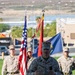 A U.S. Marine in desert MARPAT camouflage uniform is speaking at a podium holding a microphone during what appears to be a formal military ceremony outdoors. Behind him, a color guard stands at attention holding the U.S. flag, a red Marine Corps flag with streamers, and a blue unit flag. The setting appears to be a military installation, likely in a desert region based on the terrain visible in the background.