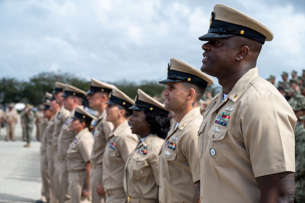A formation of United States Navy personnel in khaki service uniforms standing at attention during what appears to be a formal military ceremony or parade. The foreground features a senior enlisted sailor with multiple service ribbons and warfare devices prominently displayed. The group is photographed outdoors under a partly cloudy sky.