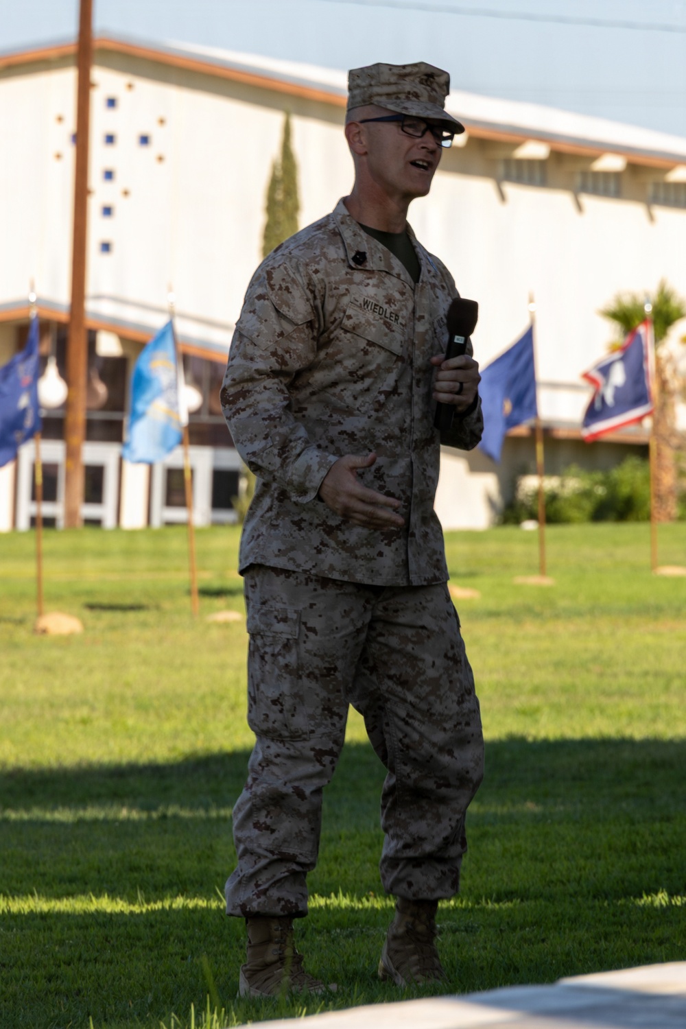 A U.S. Marine in MARPAT desert camouflage uniform is speaking into a handheld microphone on a grass field during what appears to be an official military ceremony or event. The name tape on his uniform reads 'WIEDLER' and he wears glasses and a cover (cap). Multiple state or unit flags are displayed on poles in the background.