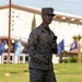 A U.S. Marine in MARPAT desert camouflage uniform is speaking into a handheld microphone on a grass field during what appears to be an official military ceremony or event. The name tape on his uniform reads 'WIEDLER' and he wears glasses and a cover (cap). Multiple state or unit flags are displayed on poles in the background.