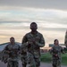 A group of U.S. Army soldiers in Operational Camouflage Pattern (OCP) uniforms are running on a gravel path during what appears to be an early morning or evening physical training exercise. The lead soldier, identified by name tape as 'AUDREY,' is running ahead of the group with visible effort. The setting appears to be a military installation with buildings and green grass visible in the background.