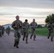 A group of U.S. Army soldiers in Operational Camouflage Pattern (OCP) uniforms are conducting a formation run along a gravel path at what appears to be a military installation. The run is taking place during early morning or evening hours based on the overcast, low-light sky conditions. A lead soldier wearing glasses and bearing a visible name tape is at the front of the formation.