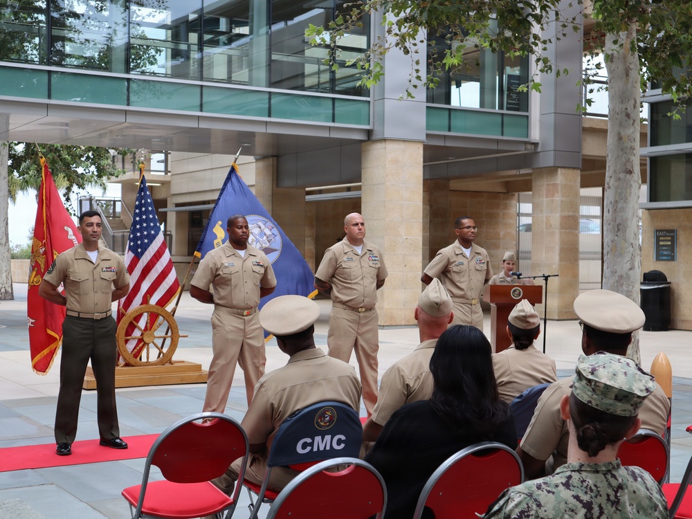 A formal military ceremony is taking place outdoors in a plaza area in front of a modern glass and stone building. Four uniformed military personnel stand at attention on a red carpet, with flags including the US flag, a blue Navy flag, and a red Marine Corps flag displayed behind them. An audience of uniformed service members is seated in folding chairs facing the standing personnel, with a podium visible in the background.