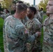 A U.S. Army officer is pinning an award or insignia onto a soldier during what appears to be an outdoor military ceremony on a grassy field. Multiple soldiers in Operational Camouflage Pattern (OCP) uniforms are standing in formation in the background. The scene suggests a formal recognition or promotion ceremony taking place at dusk or early morning.