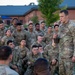A U.S. Army officer, identifiable by rank insignia and 'U.S. ARMY' tape, addresses a large group of soldiers gathered outdoors on what appears to be a military installation. The soldiers are wearing Operational Camouflage Pattern (OCP) uniforms and appear to be listening attentively during what looks like a briefing or after-action review. The setting appears to be early morning or evening based on the ambient lighting.