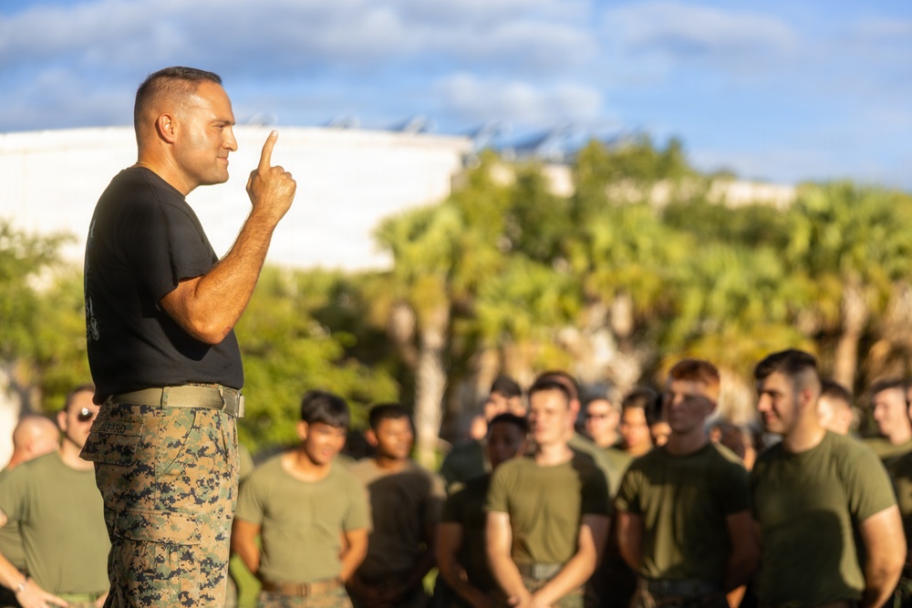 A military leader, wearing a black t-shirt and MARPAT camouflage trousers, addresses a large group of uniformed personnel outdoors. He is gesturing with one finger raised, suggesting he is making a key point during a briefing or formation. The setting appears to be a military installation with palm trees and large storage tanks visible in the background.