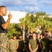 A military leader, wearing a black t-shirt and MARPAT camouflage trousers, addresses a large group of uniformed personnel outdoors. He is gesturing with one finger raised, suggesting he is making a key point during a briefing or formation. The setting appears to be a military installation with palm trees and large storage tanks visible in the background.