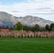 A large group of military personnel in camouflage uniforms are posed for a formal unit photograph on a green athletic field. They are arranged in multiple rows with some kneeling in front, set against a backdrop of prominent rocky mountains and trees. The setting appears to be a military installation, likely in the Colorado Springs area based on the distinctive mountain profile.
