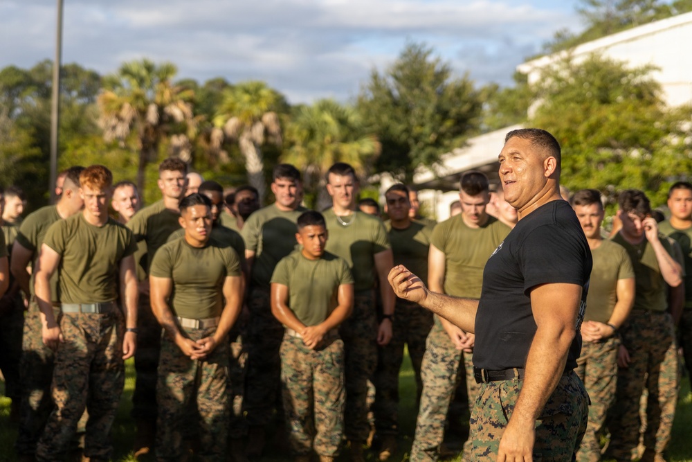 A muscular individual wearing a dark navy t-shirt and camouflage trousers addresses a large group of military personnel in green/camouflage uniforms outdoors. The setting appears to be a military base or training facility, with palm trees and a building visible in the background. The speaker appears to be in a leadership or instructional role, gesturing while speaking to the assembled group.