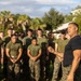 A muscular individual wearing a dark navy t-shirt and camouflage trousers addresses a large group of military personnel in green/camouflage uniforms outdoors. The setting appears to be a military base or training facility, with palm trees and a building visible in the background. The speaker appears to be in a leadership or instructional role, gesturing while speaking to the assembled group.
