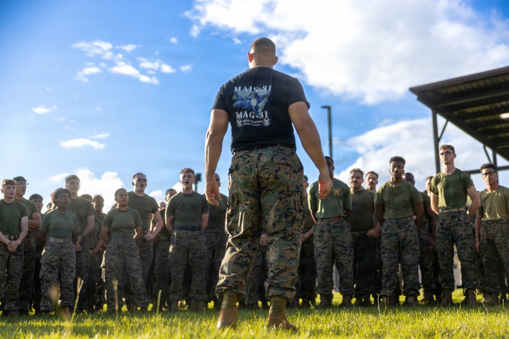 A military instructor or senior NCO wearing a black 'MALS-31 / MAG-31' t-shirt and MARPAT camouflage trousers stands with his back to the camera, addressing a large formation of U.S. Marines in olive drab PT gear and camouflage utilities. The group appears to be assembled outdoors on a grass field, likely for a physical training event, briefing, or formation. The setting suggests a U.S. Marine Corps installation.