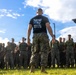 A military instructor or senior NCO wearing a black 'MALS-31 / MAG-31' t-shirt and MARPAT camouflage trousers stands with his back to the camera, addressing a large formation of U.S. Marines in olive drab PT gear and camouflage utilities. The group appears to be assembled outdoors on a grass field, likely for a physical training event, briefing, or formation. The setting suggests a U.S. Marine Corps installation.