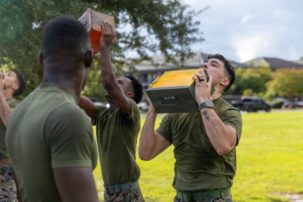 A group of military personnel in olive drab PT (physical training) uniforms are performing an exercise involving lifting or pressing heavy metal ammunition cans overhead. The activity appears to be a physical fitness training exercise conducted outdoors on a grassy field. Multiple service members are visible participating simultaneously with similar metal containers.