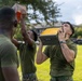 A group of military personnel in olive drab PT (physical training) uniforms are performing an exercise involving lifting or pressing heavy metal ammunition cans overhead. The activity appears to be a physical fitness training exercise conducted outdoors on a grassy field. Multiple service members are visible participating simultaneously with similar metal containers.