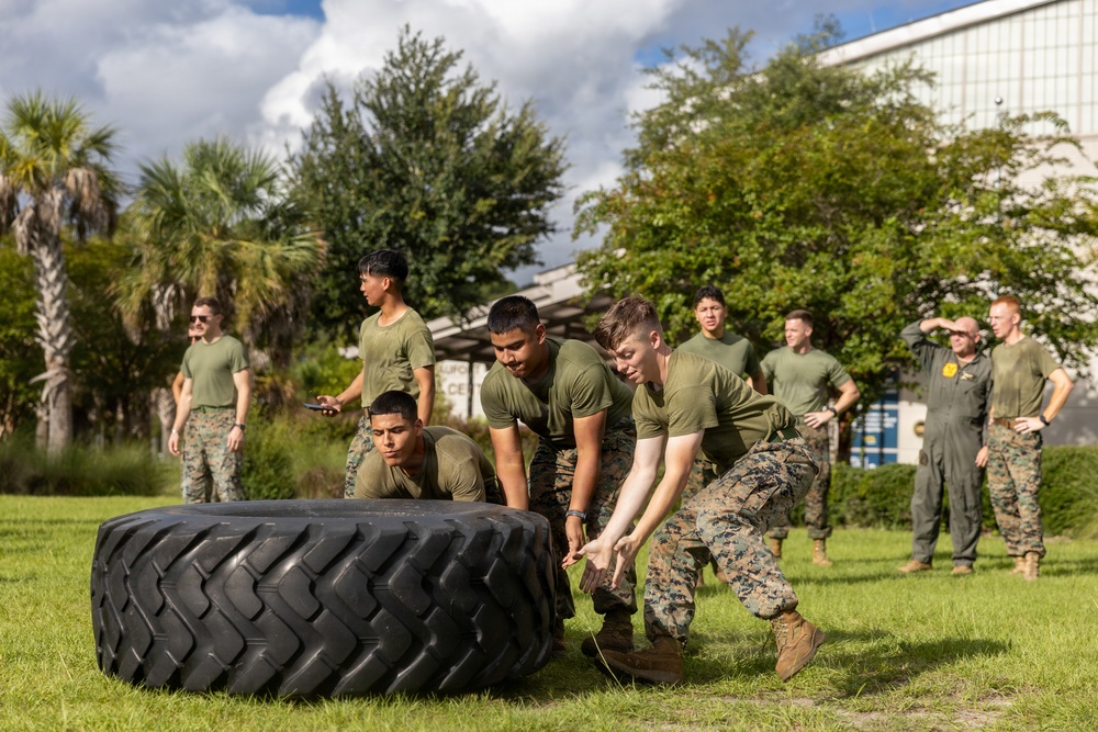 A group of military personnel in olive drab PT (physical training) uniforms are performing a tire flip exercise on a grass field. Several individuals are actively flipping a large heavy-duty tractor tire while others observe from the background. One person in a yellow flight suit is also visible among the observers, suggesting a mixed-unit training event.