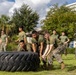 A group of military personnel in olive drab PT (physical training) uniforms are performing a tire flip exercise on a grass field. Several individuals are actively flipping a large heavy-duty tractor tire while others observe from the background. One person in a yellow flight suit is also visible among the observers, suggesting a mixed-unit training event.