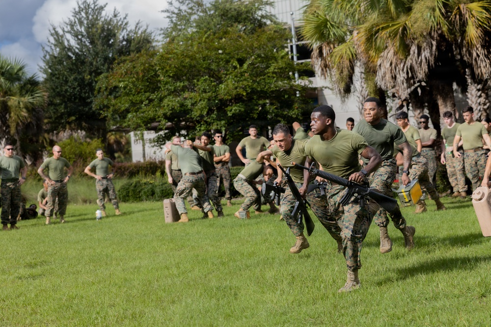 A group of military personnel, appearing to be U.S. Marines based on their distinctive woodland camouflage uniforms and olive drab t-shirts, are conducting an outdoor physical training or combat fitness exercise on a grass field. In the foreground, two Marines are carrying a rifle and what appears to be an ammunition can while running, suggesting a combat fitness relay or team challenge event. A large group of Marines observes and participates in the background, with palm trees and a building visible in the setting.