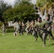 A group of military personnel, appearing to be U.S. Marines based on their distinctive woodland camouflage uniforms and olive drab t-shirts, are conducting an outdoor physical training or combat fitness exercise on a grass field. In the foreground, two Marines are carrying a rifle and what appears to be an ammunition can while running, suggesting a combat fitness relay or team challenge event. A large group of Marines observes and participates in the background, with palm trees and a building visible in the setting.