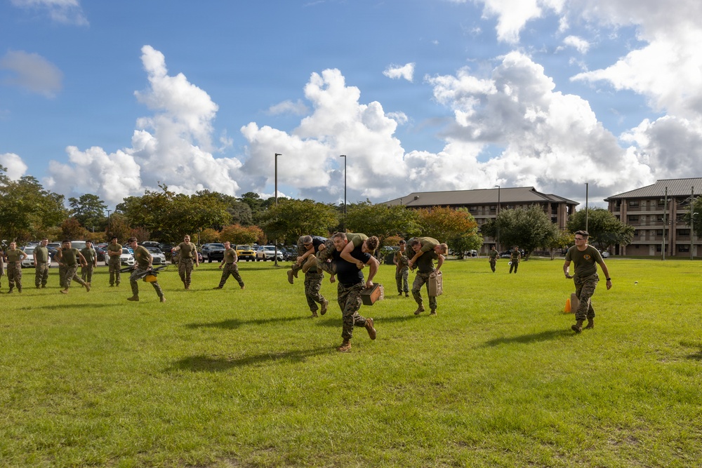A group of military personnel, appearing to be U.S. Marines based on their camouflage uniforms, are conducting a physical training exercise on a grassy field. The exercise involves carrying fellow service members on their backs while running, suggesting a combat casualty evacuation or buddy-carry drill. The activity takes place on what appears to be a military installation, with barracks-style buildings and a parking lot visible in the background.