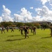 A group of military personnel, appearing to be U.S. Marines based on their camouflage uniforms, are conducting a physical training exercise on a grassy field. The exercise involves carrying fellow service members on their backs while running, suggesting a combat casualty evacuation or buddy-carry drill. The activity takes place on what appears to be a military installation, with barracks-style buildings and a parking lot visible in the background.