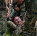 Multiple military personnel in woodland digital camouflage uniforms are engaged in a close-quarters combat or martial arts training exercise on grass. One service member wearing a black mouthguard is being held in a grappling position by another, with additional personnel surrounding them. The scene depicts an organized combatives or hand-to-hand combat training drill.