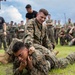 A group of military personnel, appearing to be U.S. Marines based on their MARPAT camouflage uniforms, are engaged in a martial arts or combatives training exercise outdoors on a grass field. Two Marines are grappling on the ground in the foreground while a large crowd of fellow Marines watches from the background. The atmosphere appears to be a structured training event or competition.