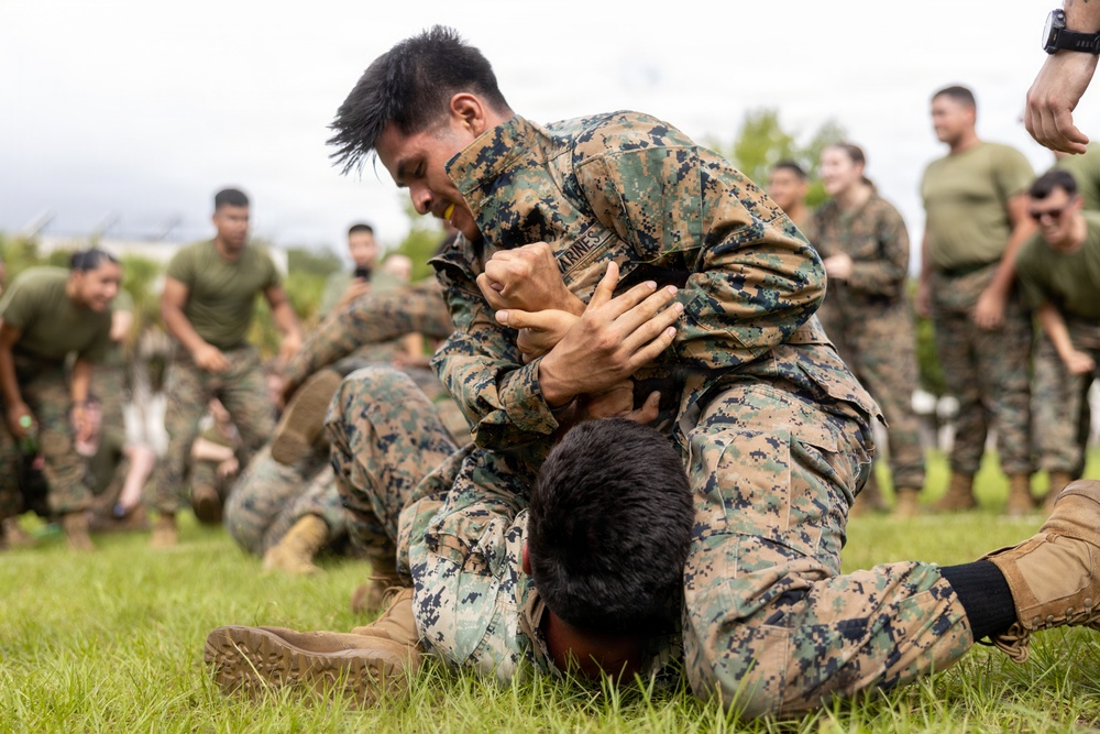 Two military personnel in camouflage uniforms are engaged in a ground grappling exercise on a grass field. The foreground combatant, wearing a uniform marked 'MARINES,' has mounted his opponent in a dominant ground position. A large group of similarly uniformed military personnel observes the sparring match in the background.