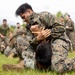 Two military personnel in camouflage uniforms are engaged in a ground grappling exercise on a grass field. The foreground combatant, wearing a uniform marked 'MARINES,' has mounted his opponent in a dominant ground position. A large group of similarly uniformed military personnel observes the sparring match in the background.