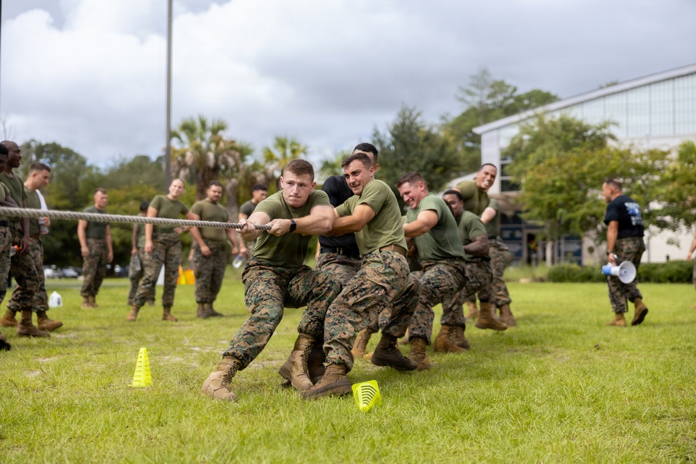 A group of military personnel in U.S. Marine Corps camouflage uniforms (MARPAT) are engaged in a tug-of-war competition on a grass field. The activity appears to be part of a unit morale or field day event, with spectators in matching uniforms watching from the sidelines. A person with a bullhorn/megaphone is visible on the right side, suggesting an organized, supervised event.