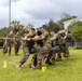 A group of military personnel in U.S. Marine Corps camouflage uniforms (MARPAT) are engaged in a tug-of-war competition on a grass field. The activity appears to be part of a unit morale or field day event, with spectators in matching uniforms watching from the sidelines. A person with a bullhorn/megaphone is visible on the right side, suggesting an organized, supervised event.