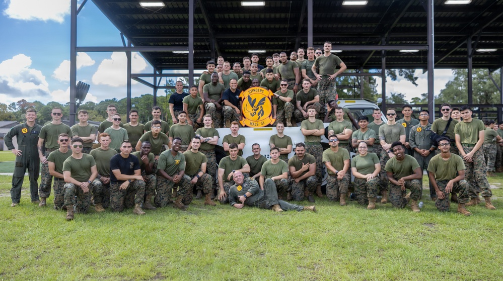 A large group of U.S. Marines poses for an official unit photograph outdoors. They are members of MALS-31 'Stingers,' as indicated by the unit patch/banner held in the center of the group. The photo appears to be taken at a military installation, likely Marine Corps Air Station Beaufort or a similar facility.