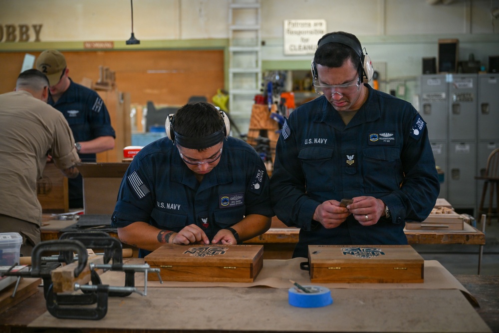 Two U.S. Navy personnel in uniform are working on wooden boxes in what appears to be a hobby or woodworking shop, both wearing ear protection and safety glasses. A third individual in tan clothing is visible in the background also working at a bench. The wooden boxes on the workbench appear to have engraved or carved designs on their lids.