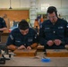 Two U.S. Navy personnel in uniform are working on wooden boxes in what appears to be a hobby or woodworking shop, both wearing ear protection and safety glasses. A third individual in tan clothing is visible in the background also working at a bench. The wooden boxes on the workbench appear to have engraved or carved designs on their lids.