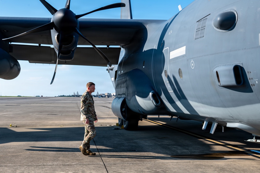 A military service member in camouflage uniform conducts a pre-flight or post-flight walk-around inspection of a large military turboprop aircraft on a flight line. The aircraft, painted in dark gray, features a prominent multi-blade propeller engine visible in the upper left. The setting appears to be a military airfield with additional aircraft visible in the background.