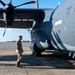 A military service member in camouflage uniform conducts a pre-flight or post-flight walk-around inspection of a large military turboprop aircraft on a flight line. The aircraft, painted in dark gray, features a prominent multi-blade propeller engine visible in the upper left. The setting appears to be a military airfield with additional aircraft visible in the background.