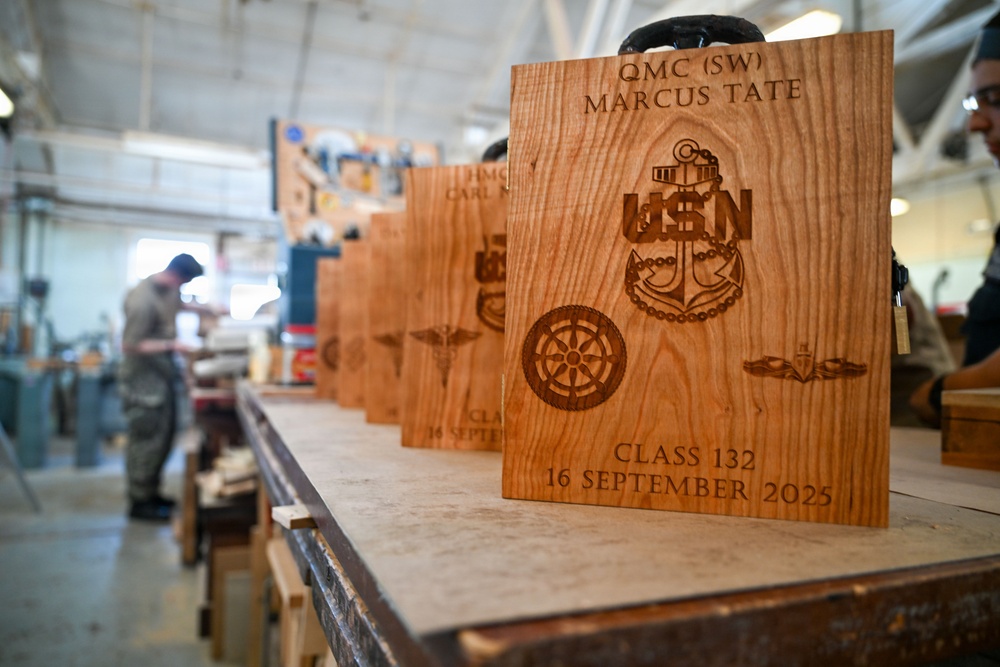 A row of laser-engraved wooden plaques are displayed on a workbench in a woodworking shop. The foremost plaque is personalized for 'QMC (SW) Marcus Tate' and commemorates 'Class 132, 16 September 2025' with U.S. Navy insignia including an anchor, USN lettering, a compass rose, and submarine warfare pins. A worker is visible in the blurred background operating machinery.