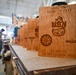 A row of laser-engraved wooden plaques are displayed on a workbench in a woodworking shop. The foremost plaque is personalized for 'QMC (SW) Marcus Tate' and commemorates 'Class 132, 16 September 2025' with U.S. Navy insignia including an anchor, USN lettering, a compass rose, and submarine warfare pins. A worker is visible in the blurred background operating machinery.