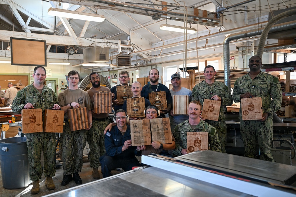 A group of approximately 13 U.S. Navy personnel pose for a group photo inside a woodworking shop, each holding custom laser-engraved wooden plaques or cutting boards. The plaques appear to feature military insignia, unit crests, flags, and personalized text. The setting is an industrial woodworking facility with lumber, machinery, and workshop equipment visible in the background.
