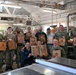 A group of approximately 13 U.S. Navy personnel pose for a group photo inside a woodworking shop, each holding custom laser-engraved wooden plaques or cutting boards. The plaques appear to feature military insignia, unit crests, flags, and personalized text. The setting is an industrial woodworking facility with lumber, machinery, and workshop equipment visible in the background.