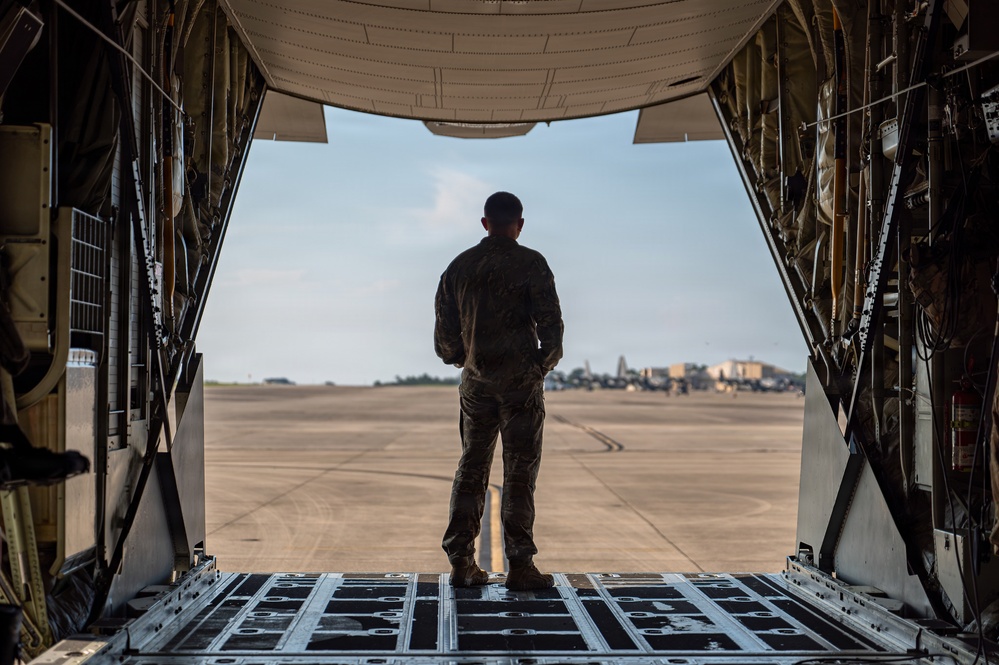 A military service member in camouflage uniform stands at the open rear cargo ramp of a large military transport aircraft, looking out onto an airfield tarmac. The perspective is from inside the aircraft looking outward, creating a dramatic framing effect with the cargo bay walls and ramp. Additional military aircraft and structures are visible in the background on the flight line.